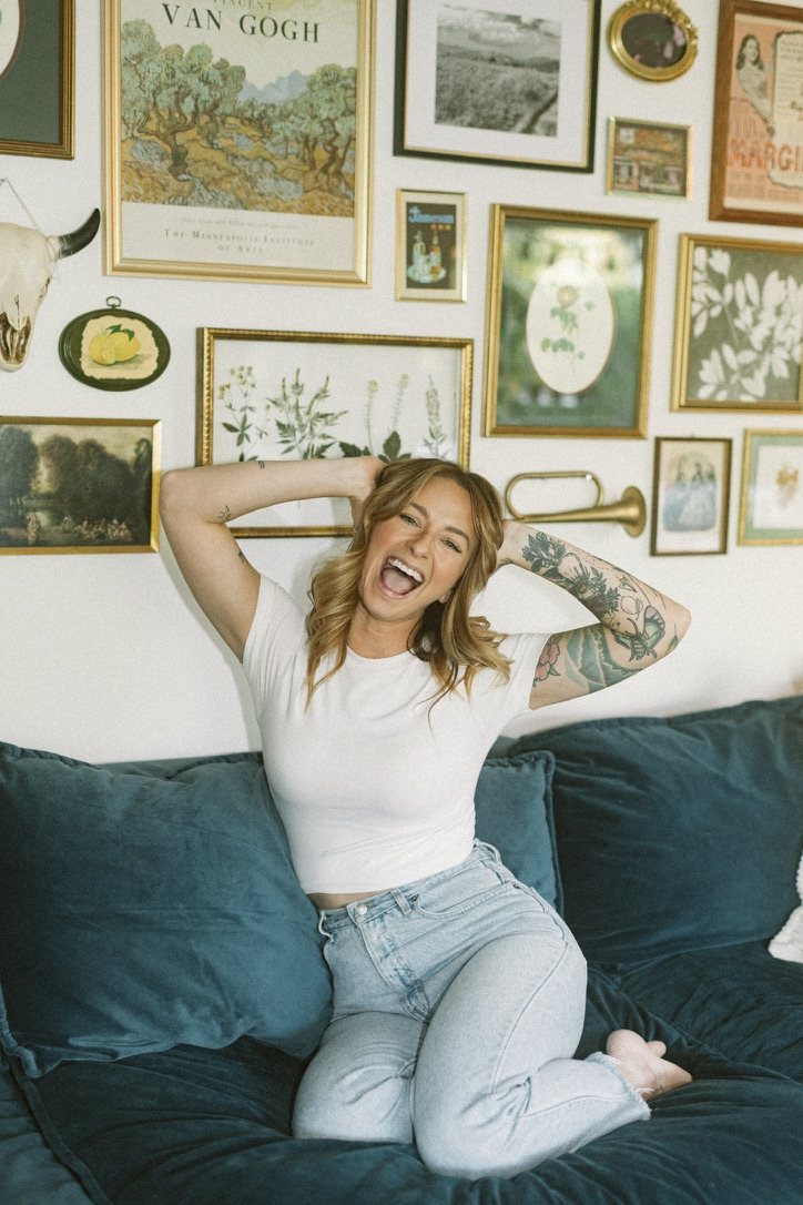 Erica laughing on a teal velvet couch in front of a gallery wall