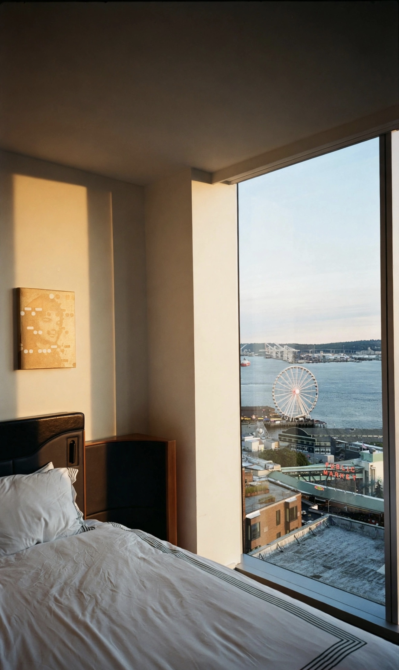 Hotel bed beside a floor-to-ceiling window looking out at the Seattle waterfront, ferris wheel and Public Market sign at golden hour